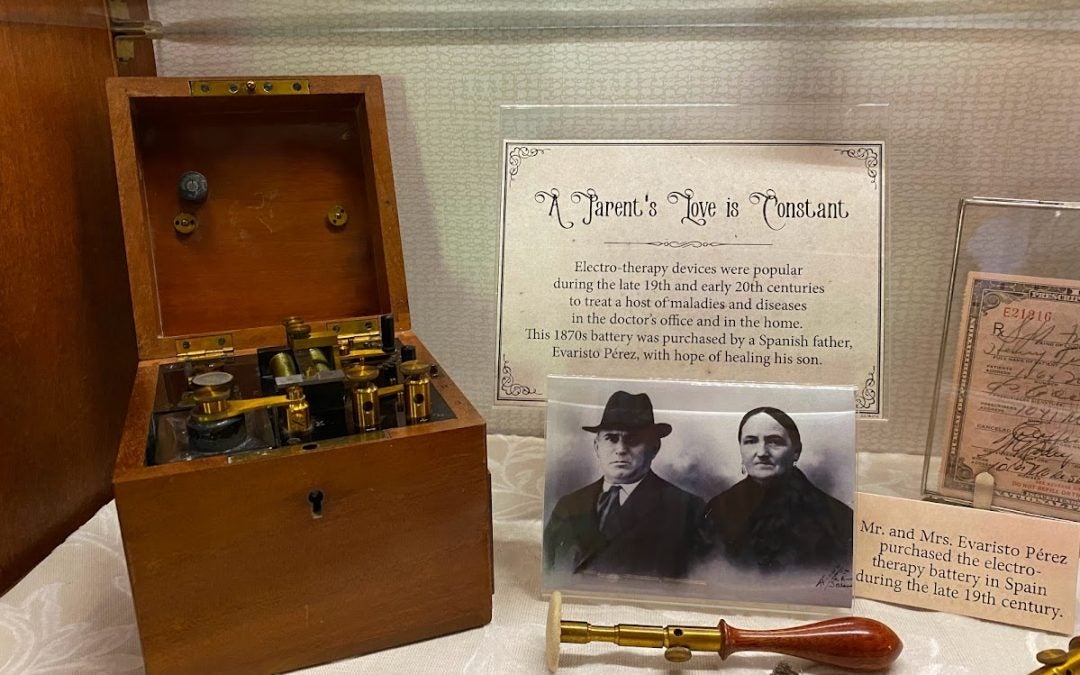 An antique medical battery box is on display with a black and white photo of an older couple and an interpretive label.